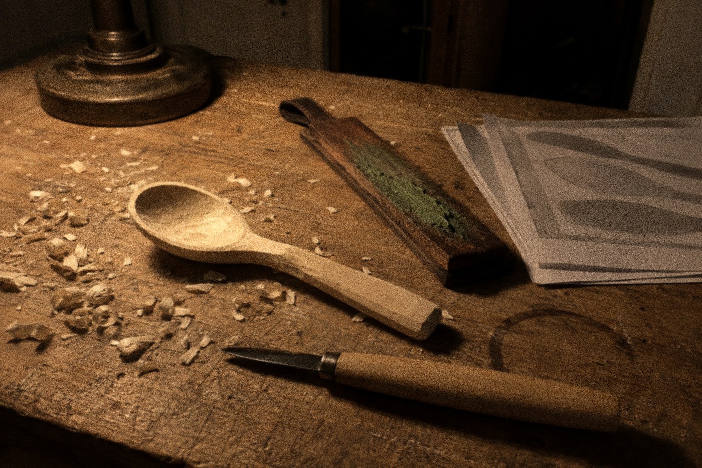 A half-carved basswood spoon on a workbench beside a leather strop and printed pattern sheets, lit by a single lamp